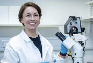 Image of speaker Jacqueline Campbell smiling, in a white lab coat standing next to a microscope.